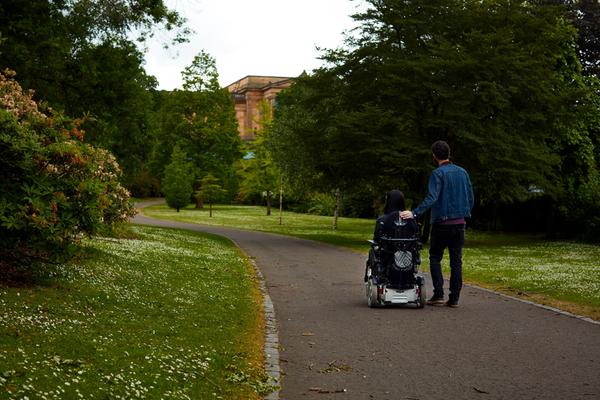 A lady in a wheelchair and her partner enjoy a walk surrounded by greenery
